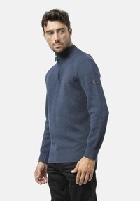 Pull à zip bleu marine en tissu tricoté, avec un col haut, des poignets côtelés et une patch logo sur la manche gauche. Design simpliste.