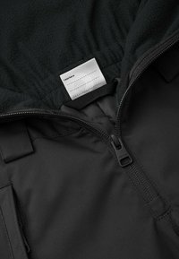 Chaqueta negra con capucha forrada de forro polar, que presenta una cremallera y una etiqueta de tela en el interior, destacando la textura del material y los detalles del diseño.