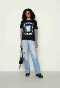 Mulher com cabelo cacheado a usar óculos de sol roxos, t-shirt gráfica preta com mangas de malha, jeans claros rasgados e sapatos pretos, segurando uma bolsa preta.