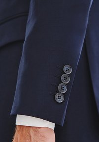 Rękaw marynarki w kolorze granatowym z czterema czarnymi guzikami, wykończony przeszyciami, z białym mankietem wystającym na zewnątrz. Gładka tekstura materiału.