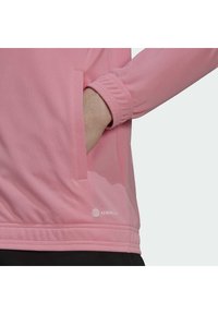 Lys rosa hettegenser laget av teksturert stoff med ribbede mansjetter, med en sidelomme og en liten logo nær kanten.