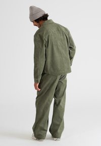 Hombre vestido con una chaqueta y pantalones de pana verde oliva, un gorro de punto beige y zapatillas blancas, de pie y mirando hacia un lado.