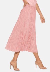 Faltender Midirock in Hellrosa, aus strukturiertem Stoff mit hoher Taille und fließender Silhouette, kombiniert mit nude-farbenen, geschnürten High Heels.