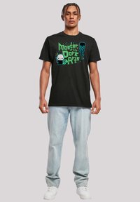 Zwarte katoenen t-shirt met een grafische print in groene en blauwe tekst en figuren, gecombineerd met lichtblauwe jeans en witte sneakers.