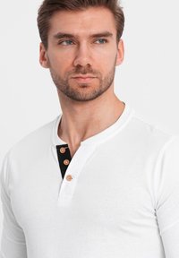 Weißes Langarm-Henley-Shirt mit schwarzem Knopfblende, drei Knopfakzenten und einer glatten Stoffoberfläche. Nahaufnahme des Ausschnitts.