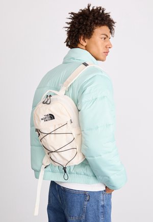 Nuori mies, jolla on vaaleansininen toppatakki ja siniset farkut sekä valkoinen The North Face -reppu yhden olan yli, sivuttain katsottuna.