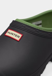 Μαύρο λαστιχένιο κλογκ με πράσινη επένδυση και ραβδωτή περιφέρεια, με ορθογώνιο σημείο λογότυπου "HUNTER" σε κόκκινο και άσπρο στο μπροστινό μέρος.