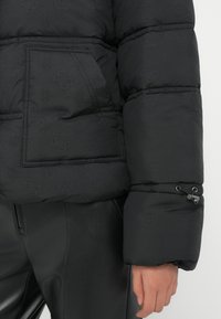 Zwarte pufferjack met een gestructureerd patroon, oversized mouwen en een zijzak met een zichtbare trekkoorddetail.