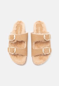 Barna műszőrmével borított papucsok, két állítható pánttal és arany csattal. Texturált felsőrész és puha bélés, lapos talp enyhe peremmel.