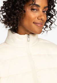 Ljusbeige zip-up jacka med hög hals, horisontell sömnad och slät textur. Har en metalldragkedja med ett hjärta som accent.