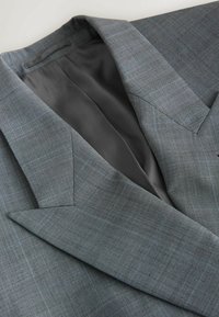 Veste de costume à carreaux gris avec revers crantés, posée à plat, montrant une doublure intérieure noire lisse et des coutures détaillées.