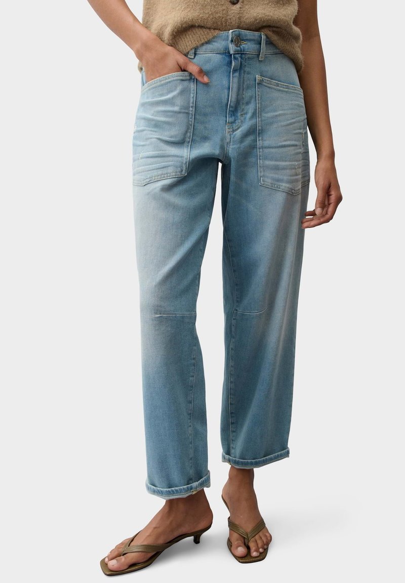Light blue high-waist jeans with large front pockets, rolled cuffs, worn with beige flip-flop heels and a brown sweater partially visible.