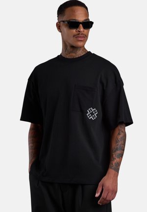 Mann trägt ein schwarzes oversized T-Shirt mit Fronttasche und weißem geometrischem Design, schwarze Sonnenbrille und tätowierte Arme, schaut nach links.