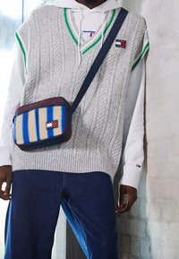 Graue Strickweste mit Zopfmuster und grünen Akzenten über einem weißen Hoodie. Das Model trägt marineblaue Jeans und eine mehrfarbige Umhängetasche mit horizontalen Streifen.