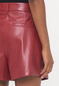Shorts rossi in pelle con una texture liscia e lucida. Presentano una vita alta e una tasca sul retro. L'orlo è leggermente svasato.