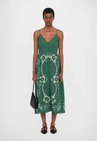 Πράσινο midi φόρεμα με λεπτές τιράντες, V neckline, floral κεντήματα, δαντελένιες λεπτομέρειες και άνετη εφαρμογή. Φορεμένο με μια μαύρη πλεκτή τσάντα.