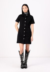Vestuário preto tipo vestido com mangas curtas, botões frontais e uma cintura definida. Usado com botas de cowboy pretas com costura decorativa branca.