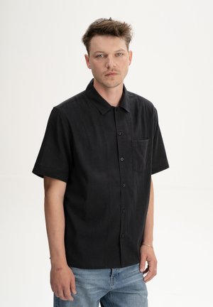 Junger Mann mit kurzen braunen Haaren, der ein schwarzes, kurzärmeliges Hemd mit Knöpfen und hellblaue Jeans trägt, vor einem schlichten hellen Hintergrund stehend.