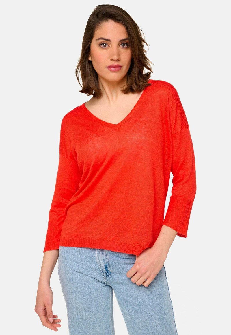 Pull-over à col en V, ample, de couleur rouge vif, fabriqué dans un tissu léger. Doté de manches trois-quarts et de poignet côtelé. Associé à un jean bleu.