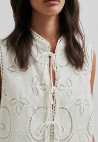 Weiße ärmellose Bluse mit gestickten Blumenmustern, gewelltem Kragen, Spitzenbesatz und einer Bindeschließe am Ausschnitt. Strukturierter Stoff.