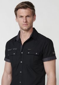 Camisa negra de manga corta hecha de algodón, con un cuello interior en contraste, dos bolsillos en el pecho y detalles de costura decorativa.