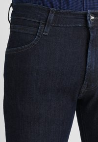 Mörkblå denimjeans med en slät yta, femfickdesign och en märkestagg i midjan. Har subtila sy detaljer.