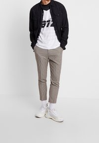 Černá košile na knoflíky přes bílé tričko s potiskem, doplněná světle hnědými kostkovanými kalhotami a bílými teniskami. Má kapsy a zúžené nohavice.