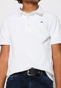 Ein Kind trägt ein weißes Polohemd mit kurzen Ärmeln und dunkle Jeans mit einem schwarzen Gürtel und steht vor einem schlichten Hintergrund.