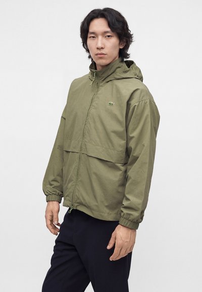 Homme portant une veste à capuche vert olive avec fermeture éclair et un pantalon noir, debout devant un fond blanc uni.