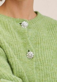 Grüner gerippter Strickpullover mit zwei dekorativen, blumenförmigen Broschen mit klaren Strasssteinen am Ausschnitt.