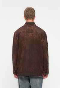 Oversized brown denim jacket with a textured finish, button cuffs, and a back label patch. Visible stitching details and relaxed fit.
