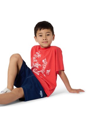 Garçon assis par terre avec une jambe repliée, portant un t-shirt rouge avec un motif graphique, un short bleu marine et des baskets blanches, sur fond blanc.