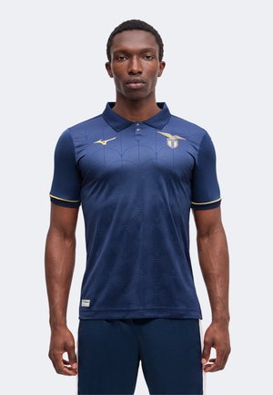 Man draagt een marineblauw poloshirt met patroon, gouden logo's en bies, staat met ontspannen armen tegen een effen lichte achtergrond.
