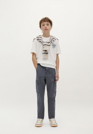 T-shirt bianca con una toppa, pantaloni cargo grigi con tasche laterali, sneakers beige e grigie, e un maglione a righe beige annodato intorno alle spalle.