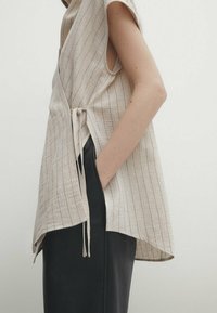 Top avvolgente beige senza maniche realizzato in un tessuto testurizzato con sottili righe verticali, dotato di un dettaglio con fiocco laterale e una vestibilità ampia. Abbinato a pantaloni neri.