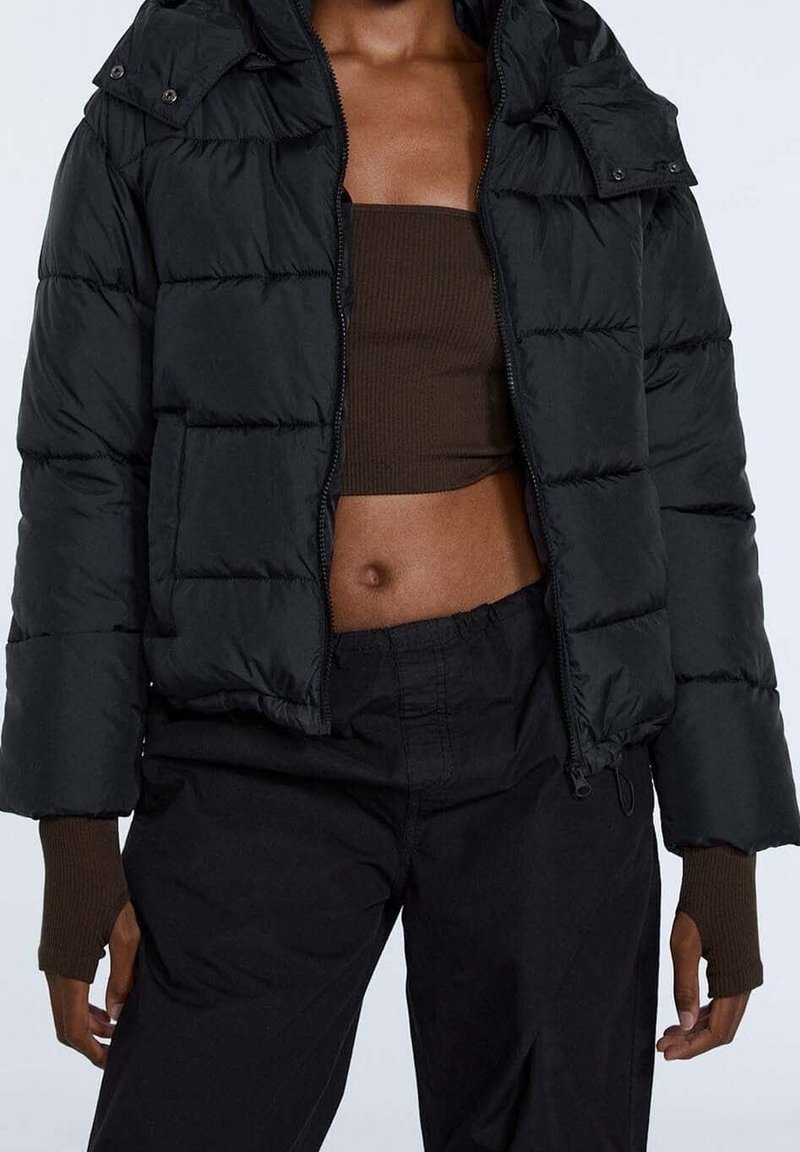 Personne portant une veste matelassée noire par-dessus un crop top marron et un pantalon noir, avec des gants marron sans doigts visibles sous les manches de la veste.