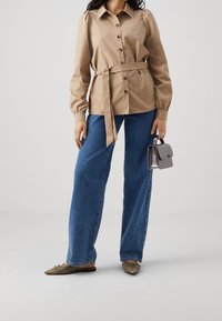 Modellen står i en stilren beige blus med bälte i midjan, kombinerad med vida blå jeans och olivgröna lägenheter, medan hon håller en liten handväska.