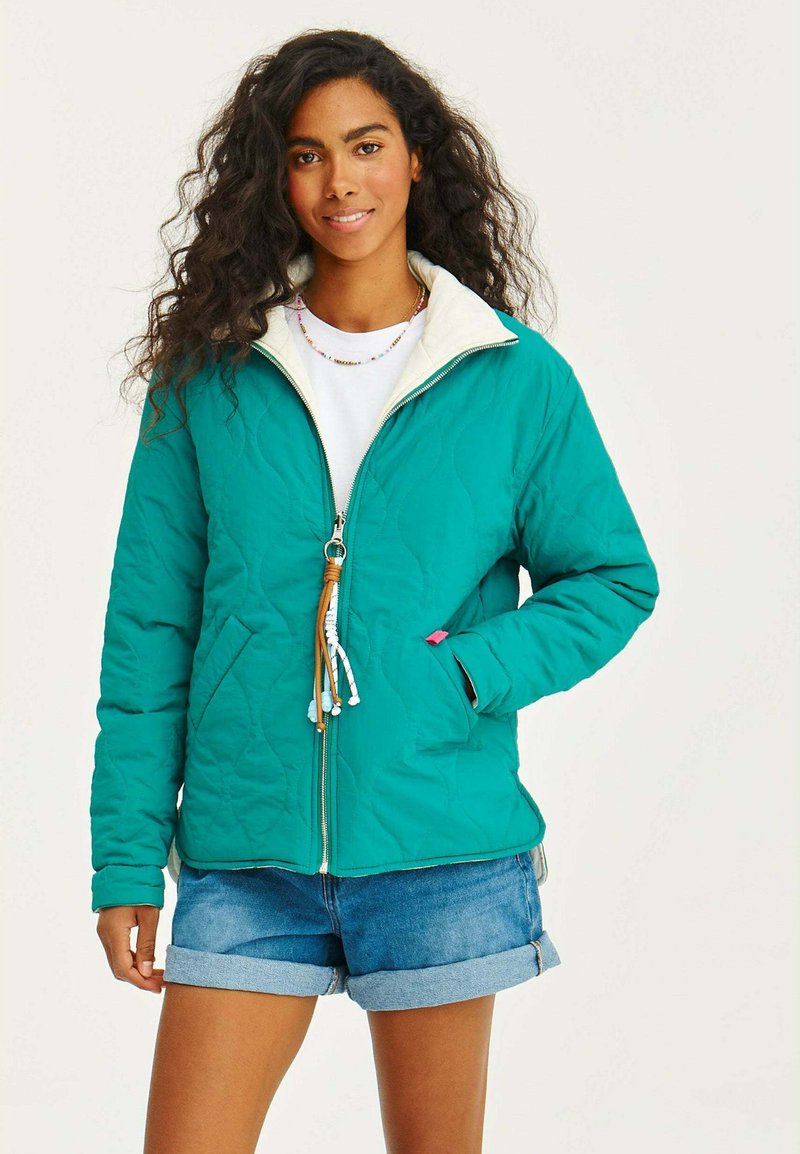 Zelená prešívaná bunda s bielym golierom, predným zipsom a detailmi s viazaním. Nosená s denimovými šortkami, predvádzajúca ležérny outfit.