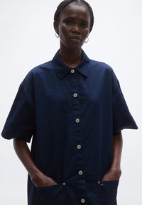 Tengerészkék rövid ujjú ing hegyes gallérral, elülső gombokkal és felvarrt zsebekkel. Simább anyagból készült, laza fazonú.