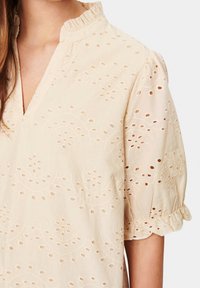 Beige puuvillapaita, jossa on röyhelö kaula-aukko, lyhyet puffihihat ja monimutkaiset brodeeratut kuvioinnit. Sisältää reikäleikkauksia ympäriinsä.