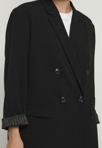 Černý dvouřadý blazer s texturovaným, plisovaným detailem na manžetách. Vybaven zaoblenými klopami a hladkou texturou tkaniny.