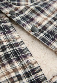Ruud kootud flanellkangas, mis sisaldab toone kreemi, musta ja pruuni, metallist nööpide ja pehme tekstureeritud fliiskattega.