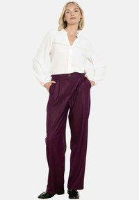 Naine seisab käed taskus, seljas valge pluus ja kõrge vöökohaga laiad lillad püksid mustade kingadega.