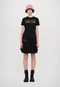 Črna majica s kratkimi rokavi z metalnim logotipom "VERSACE JEANS COUTURE", v kombinaciji s črno gubano krilo in črnimi vojaškimi čevlji. Roza kapica na glavi.