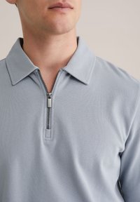 Light blue, textured polo with a zippered collar. Features a traditional collar design, high-quality fabric, and a subtle sheen.