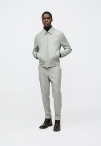 Siva jakna na pruge i odgovarajuće hlače, s ovratnikom, patentnim zatvaračem sprijeda i crnim kožnim čizmama. Glatka tekstura i kroj po mjeri.