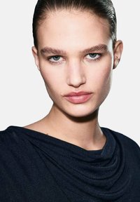 Close-up van een gezicht met een gladde huid, lichtblauwe ogen en volle lippen. Het model draagt een donkere, gedrapeerde top met een zachte textuur.