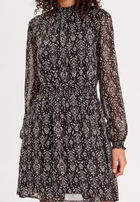 Robe noire avec manches longues transparentes, taille élastique et motifs floraux crème. Col montant, détails froncés, forme fluide.