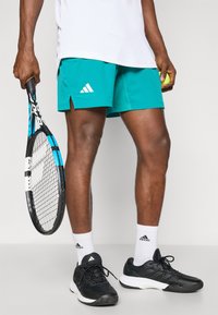 Teal sportieve shorts met wit Adidas-logo, gecombineerd met zwarte tennisschoenen, witte sokken en een tennisracket. Glad materiaal, sportief ontwerp.