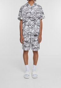 Ensemble chemise à manches courtes blanche et short, avec un motif abstrait en lignes noires, fabriqué en tissu léger, accompagné de sandales blanches à enfiler.